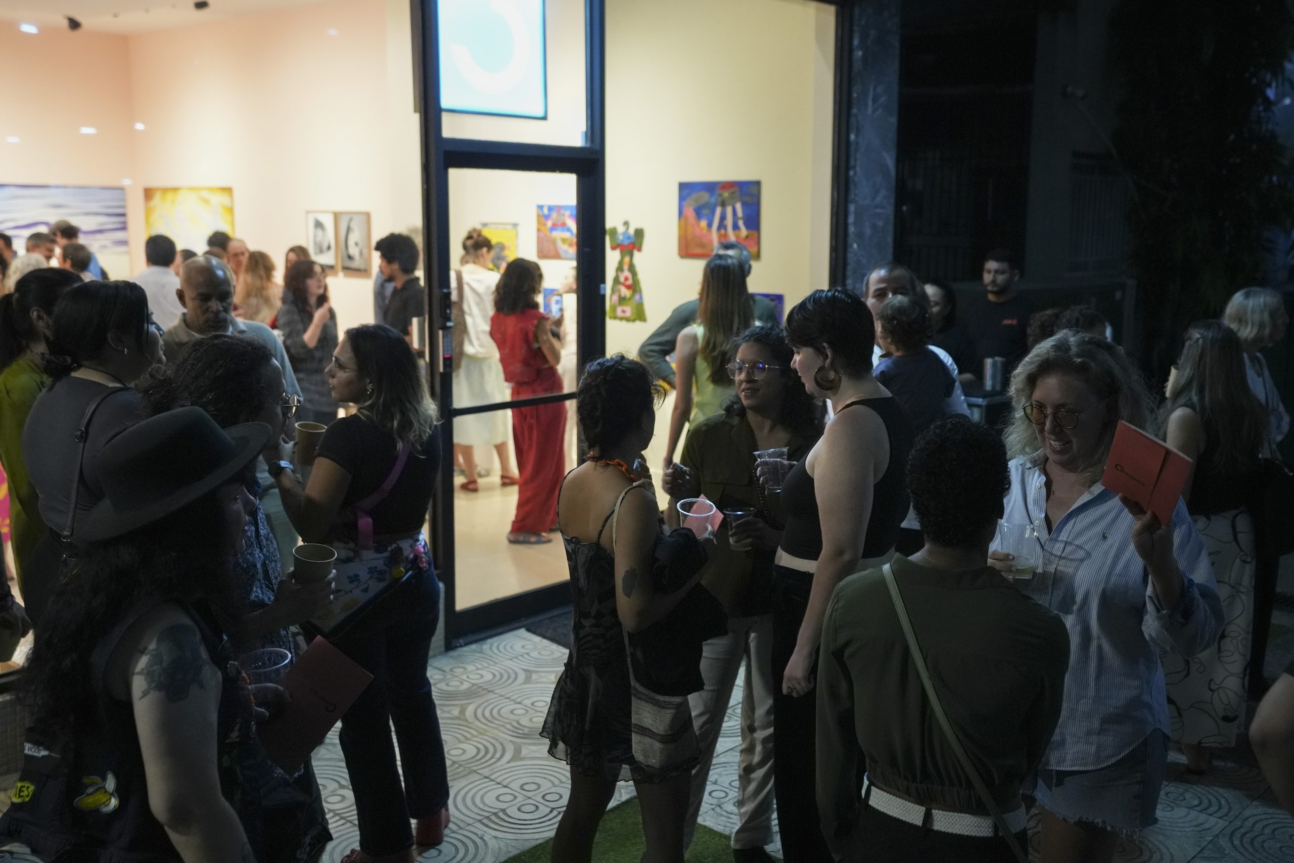 Asistentes a una inauguración nocturna conversan con vasos en la mano frente a la entrada de Arteconsult, mientras al fondo, dentro de la galería iluminada, otras personas recorren la exposición.