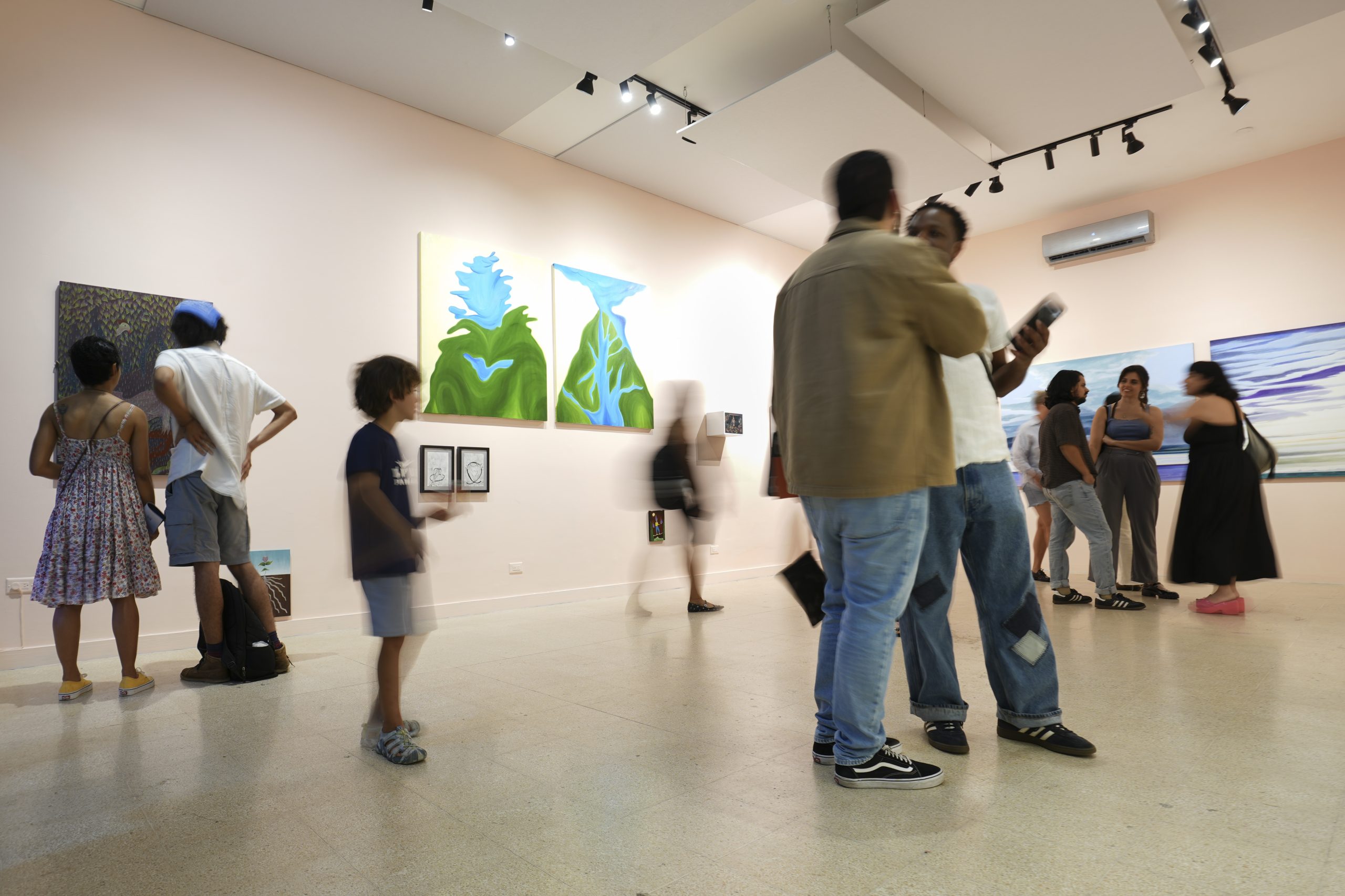 Visitantes conversan y recorren la sala principal de Arteconsult, de paredes rosa pálido y luces de riel, con pinturas de paisajes en verde y azul y obras de formato pequeño enmarcadas.