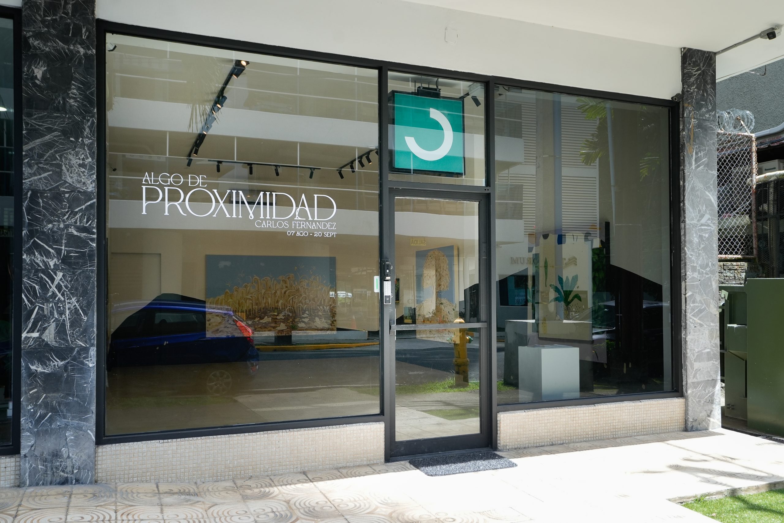 Fachada de vidrio de Arteconsult con el título de la exposición «Algo de proximidad», de Carlos Fernández, del 7 de agosto al 20 de septiembre, escrito en vinilo blanco sobre la ventana, junto al letrero verde con el logotipo de la galería.