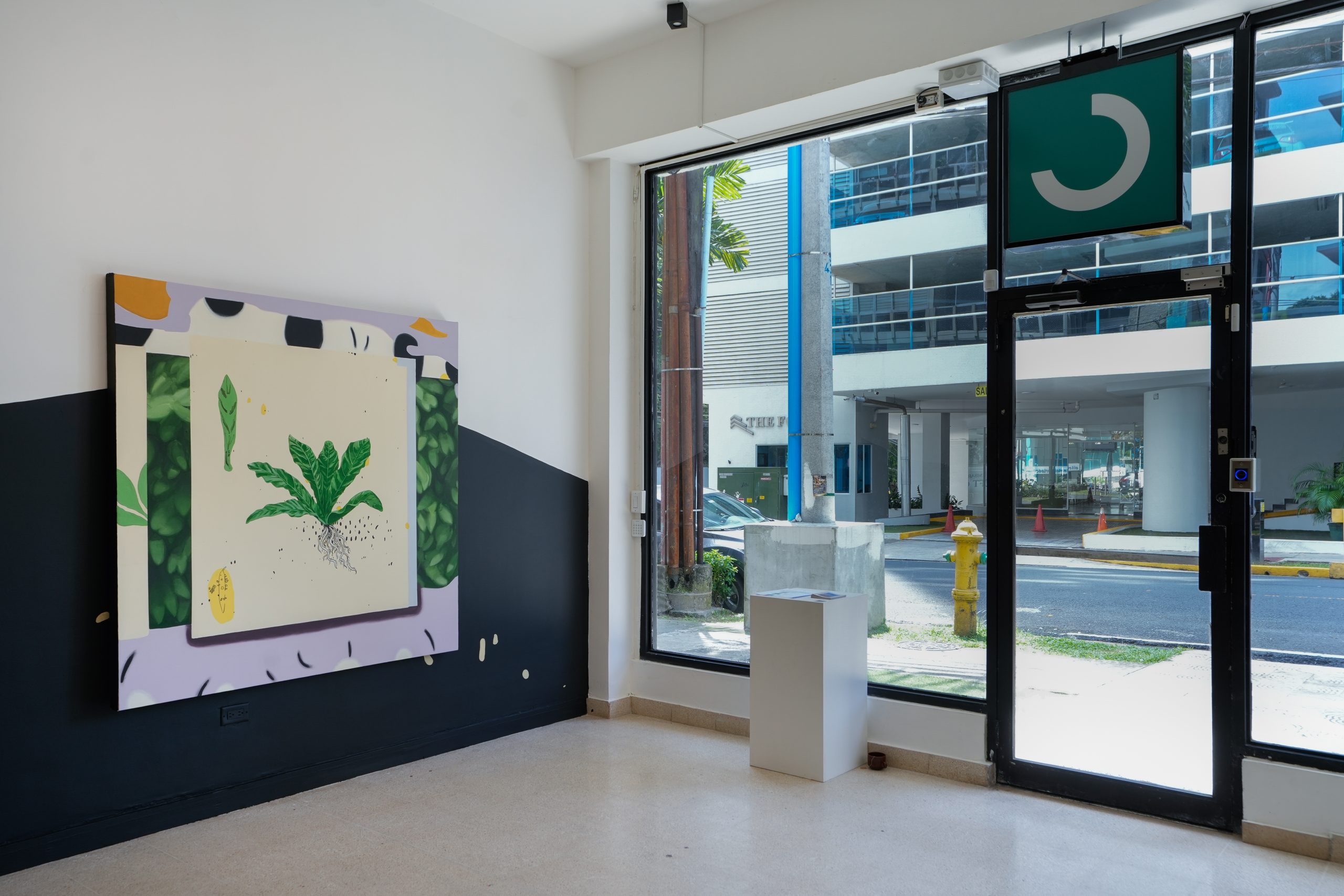 Interior de Arteconsult con una pintura de gran formato de una planta verde sobre fondo crema, colgada en una pared mitad blanca y mitad azul oscuro; junto a un pedestal blanco, el gran ventanal deja ver la calle.